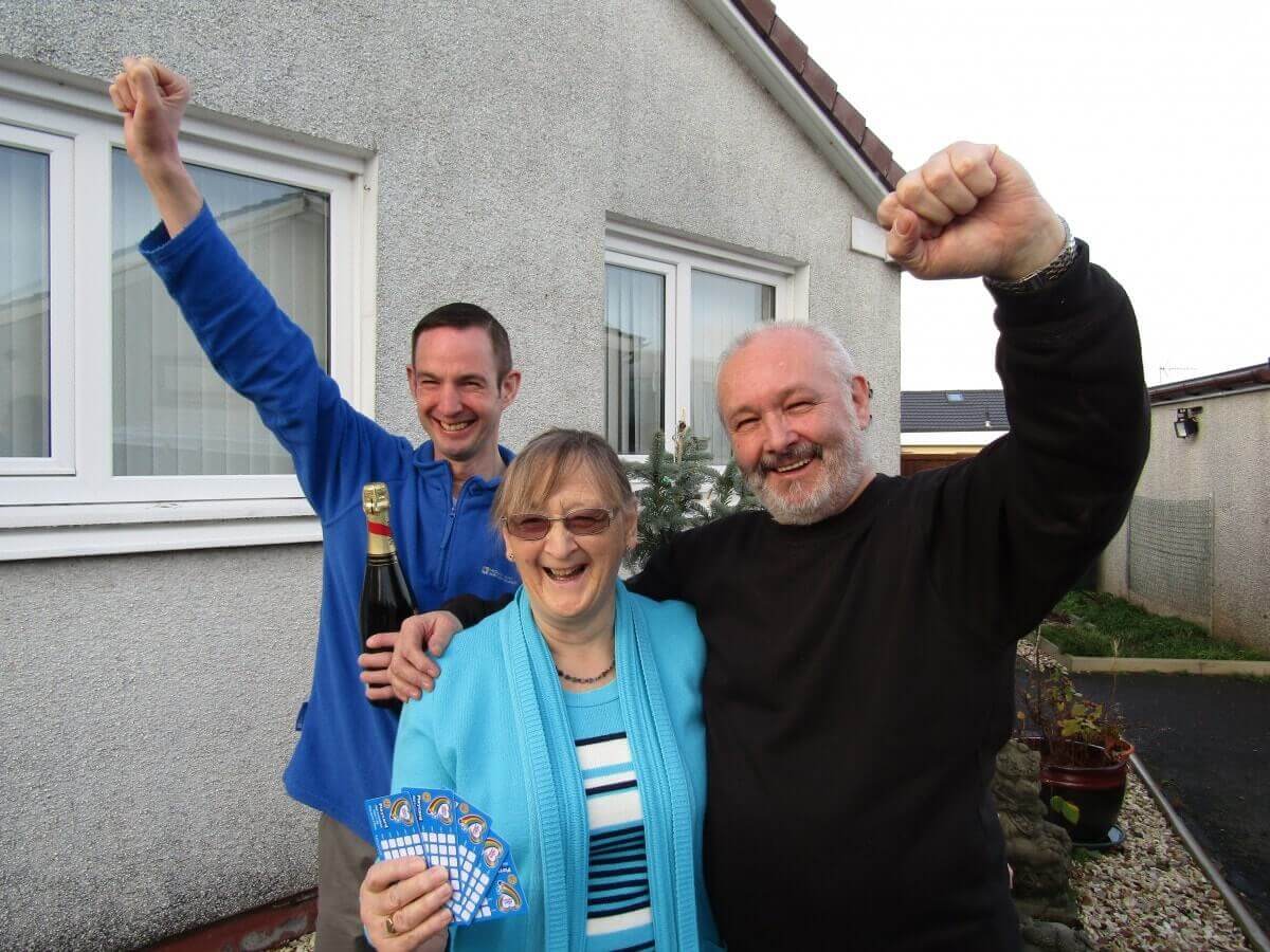 A retirement age couple and their adult son pump their fists in the air, while one holds up five scratch cards.