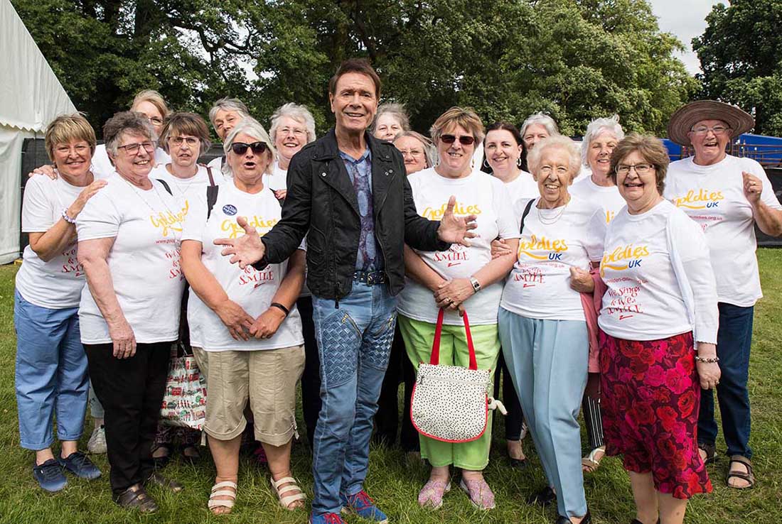 A smiling man wearing a stylish black jacket and jeans stands with a group of older women wearing matching "Goldies" tees.