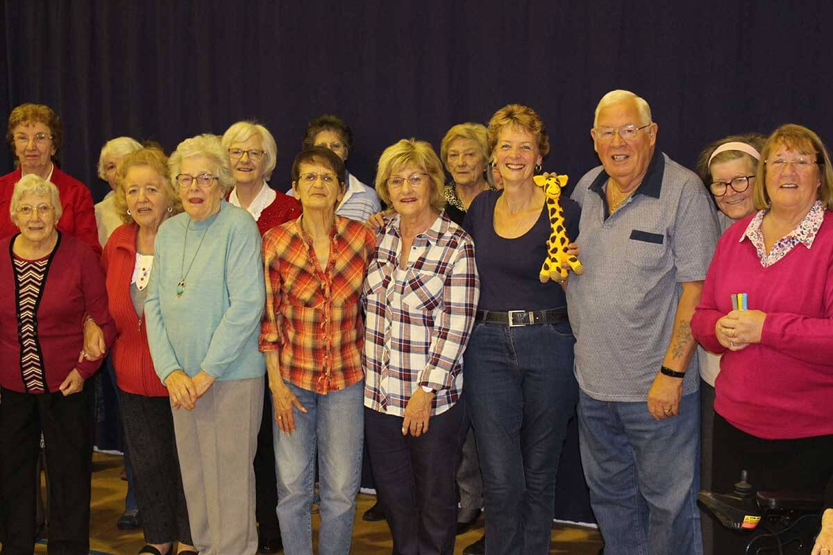 A large group of smiling senior citizens stand in front of a dark blue stage curtain.