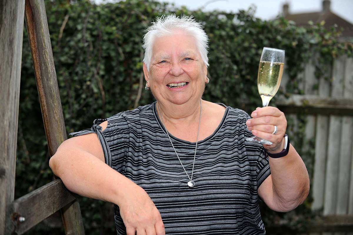 A smiling elderly woman with short white hair leans against a wooden pole, holding up a champagne glass.
