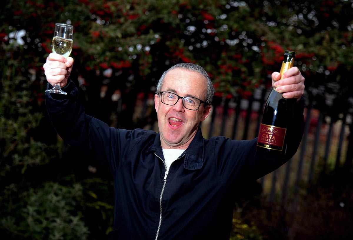 A jubliant man with glasses in a dark blue windbreaker holds up a champagne bottle in one hand and a glass in the other.