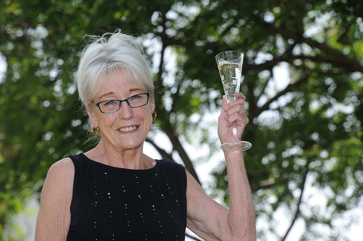 A smiling elderly white woman with white hair and glasses in a black evening dress holds up a champagne flute.